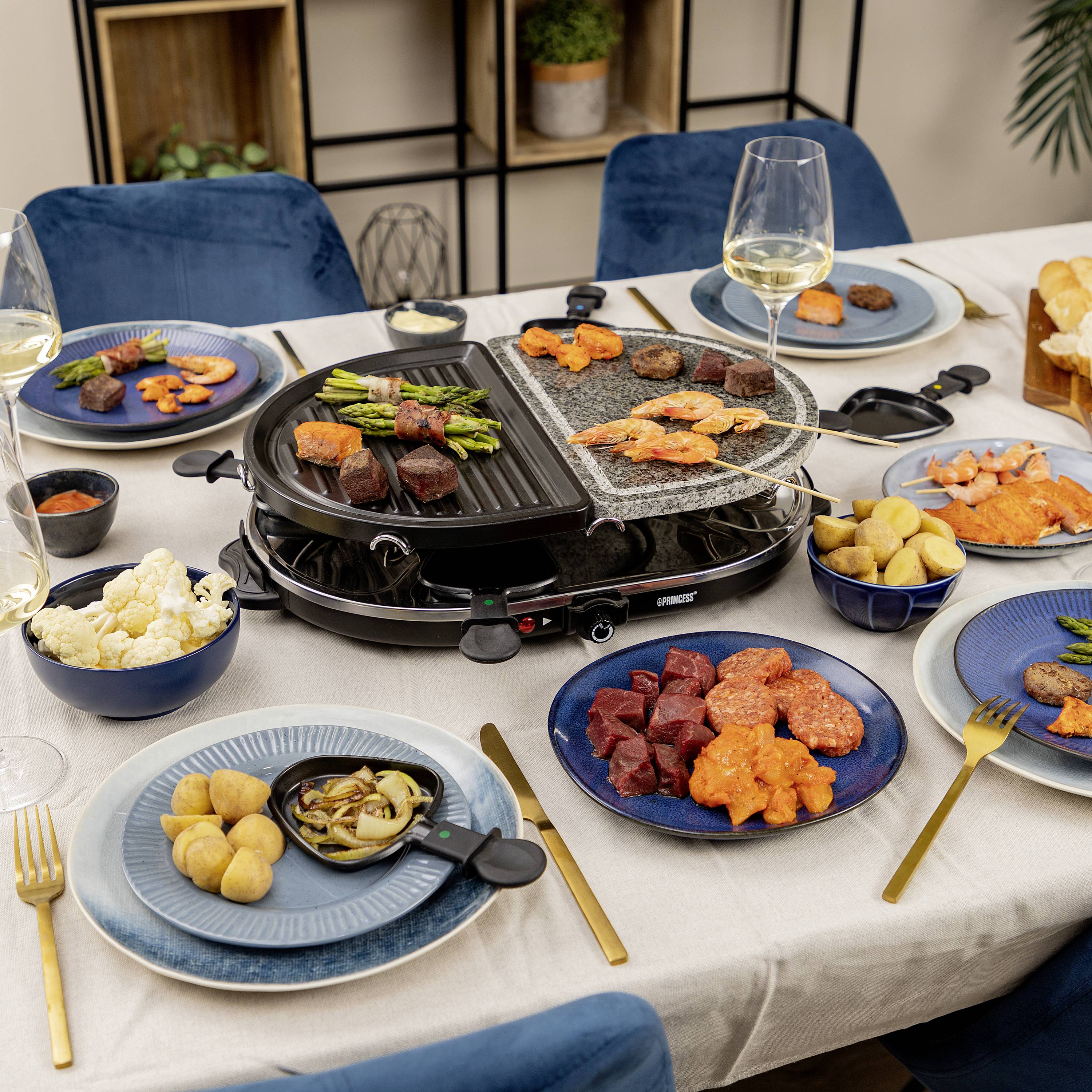 Ein Tisch mit einem Raclette-Grill, umgeben von verschiedenen Tellern mit Fleisch, Gemüse, Brot und Dips. Zwei Gläser Weißwein stehen daneben.
