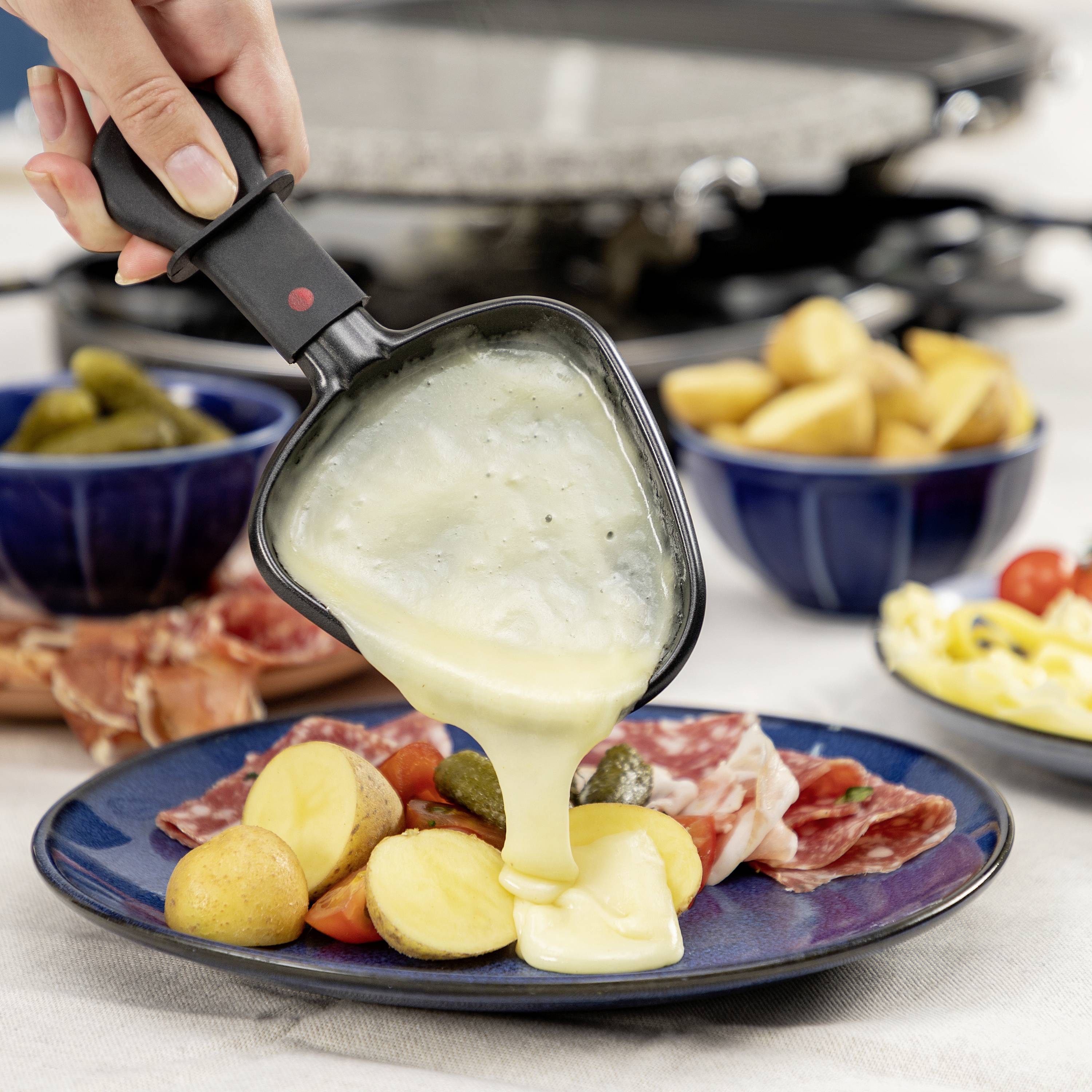 Eine Hand gießt geschmolzenen Käse aus einer Raclette-Pfanne über Kartoffeln, Gewürzgurken und Aufschnitt auf einem Teller.