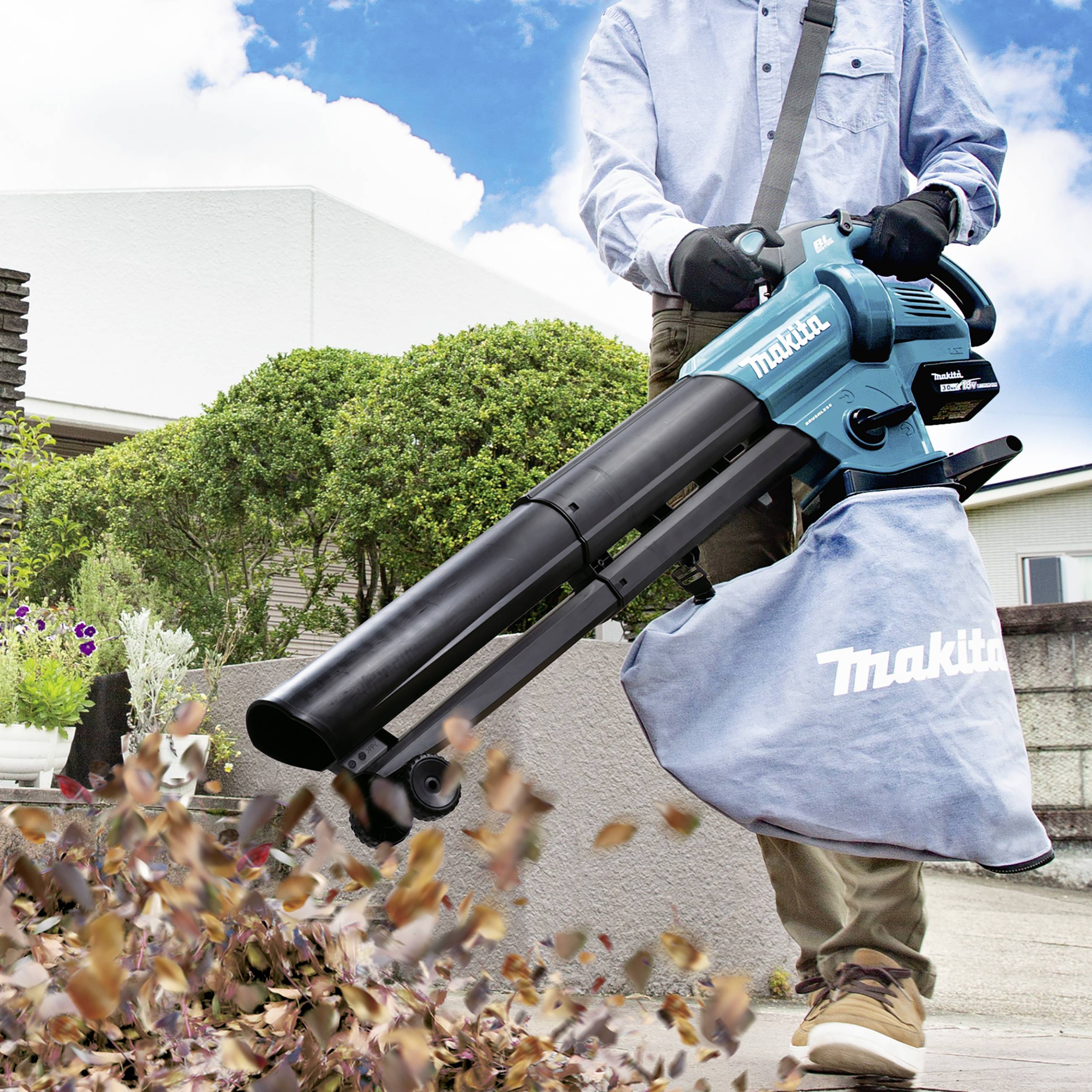 Person bläst Laub mit einem Laubbläser in einem Garten weg. Die Maschine ist blau und wird mit einem Sammelbeutel verwendet.
