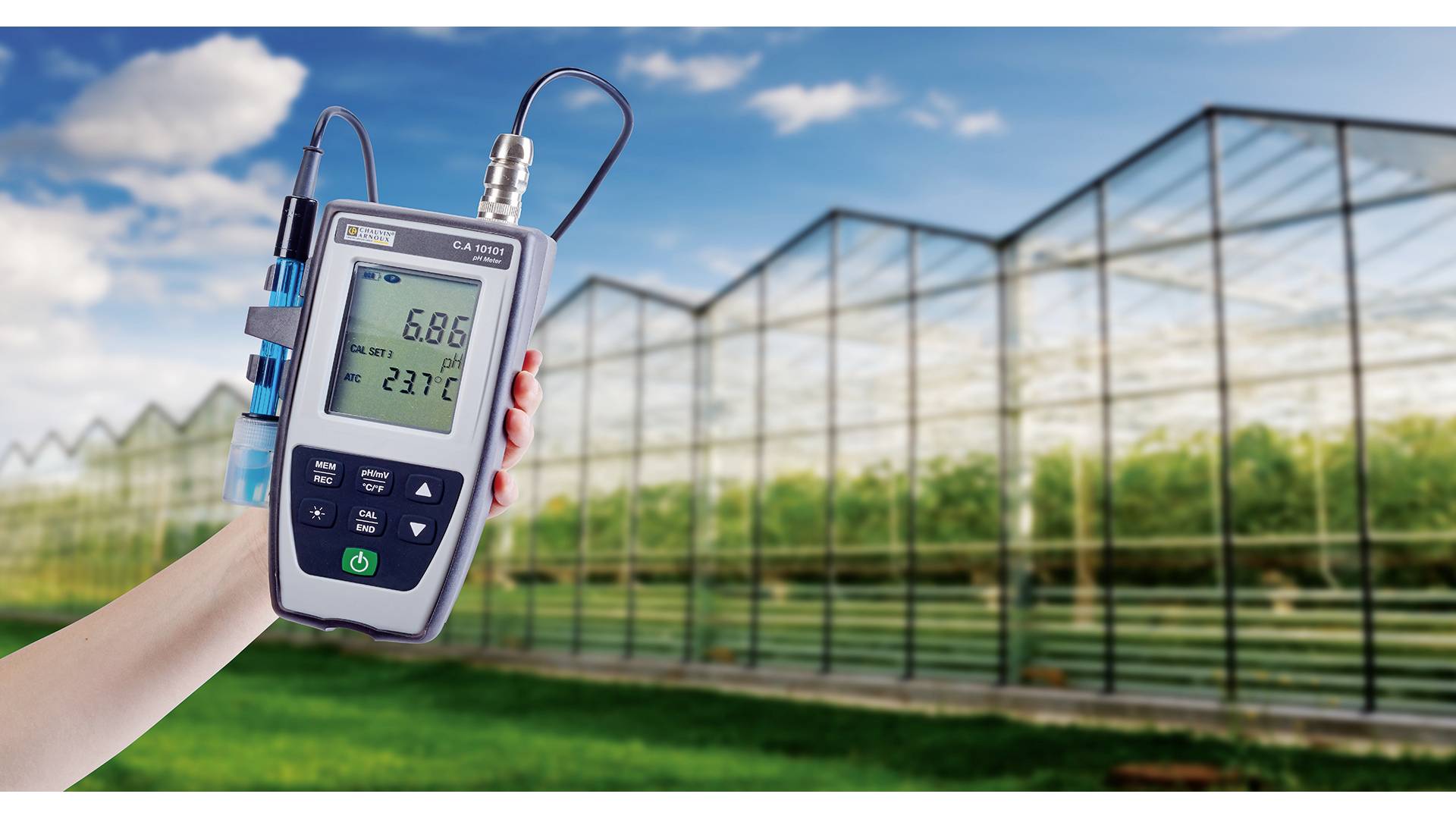 Ein tragbares Messgerät mit Display zeigt '6,86' und '23,7°C', gehalten vor einem Gewächshaus.