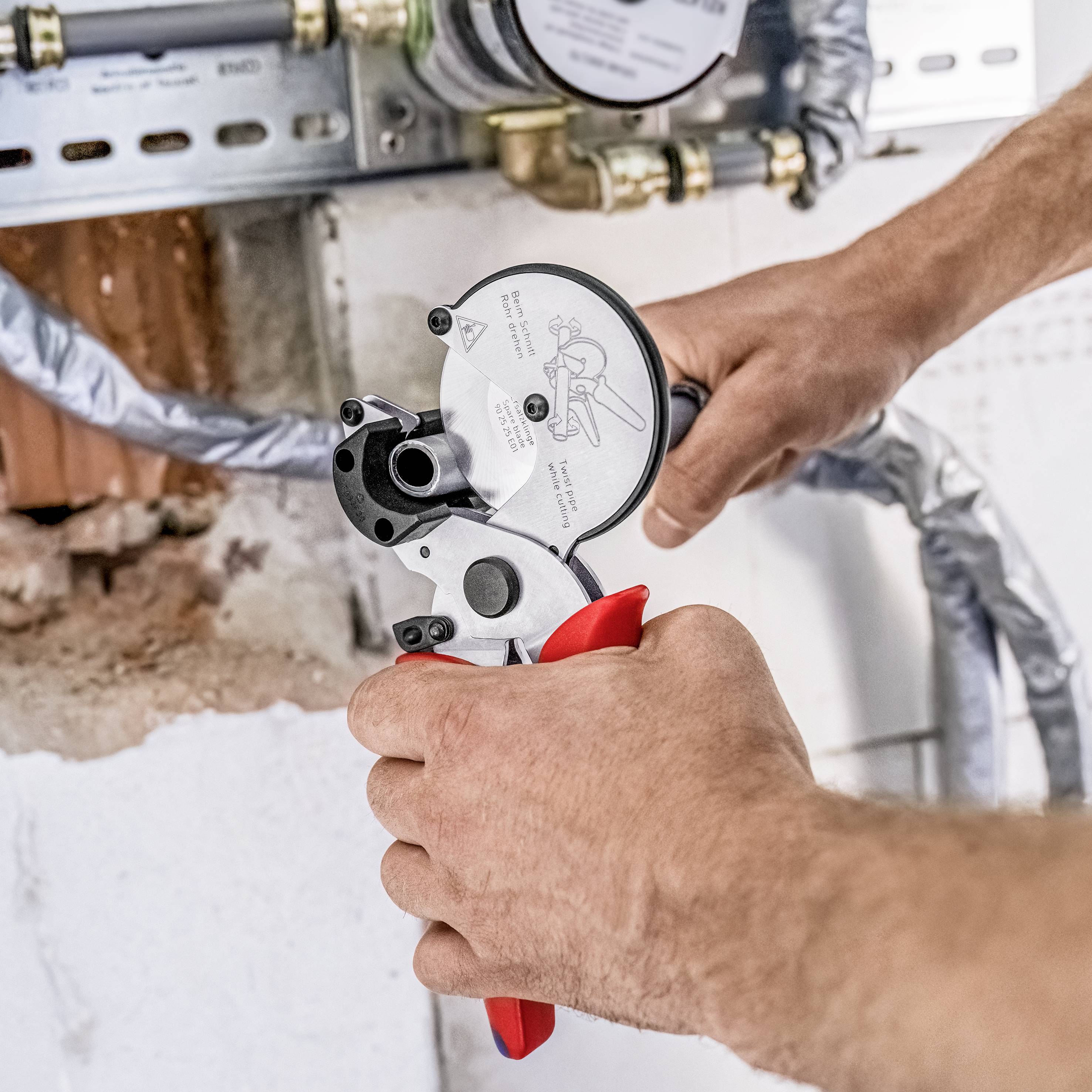 Eine Person benutzt eine Rohrpresszange, um ein Metallrohr an der Wand zu befestigen. Im Hintergrund sind Sanitärinstallationen sichtbar.