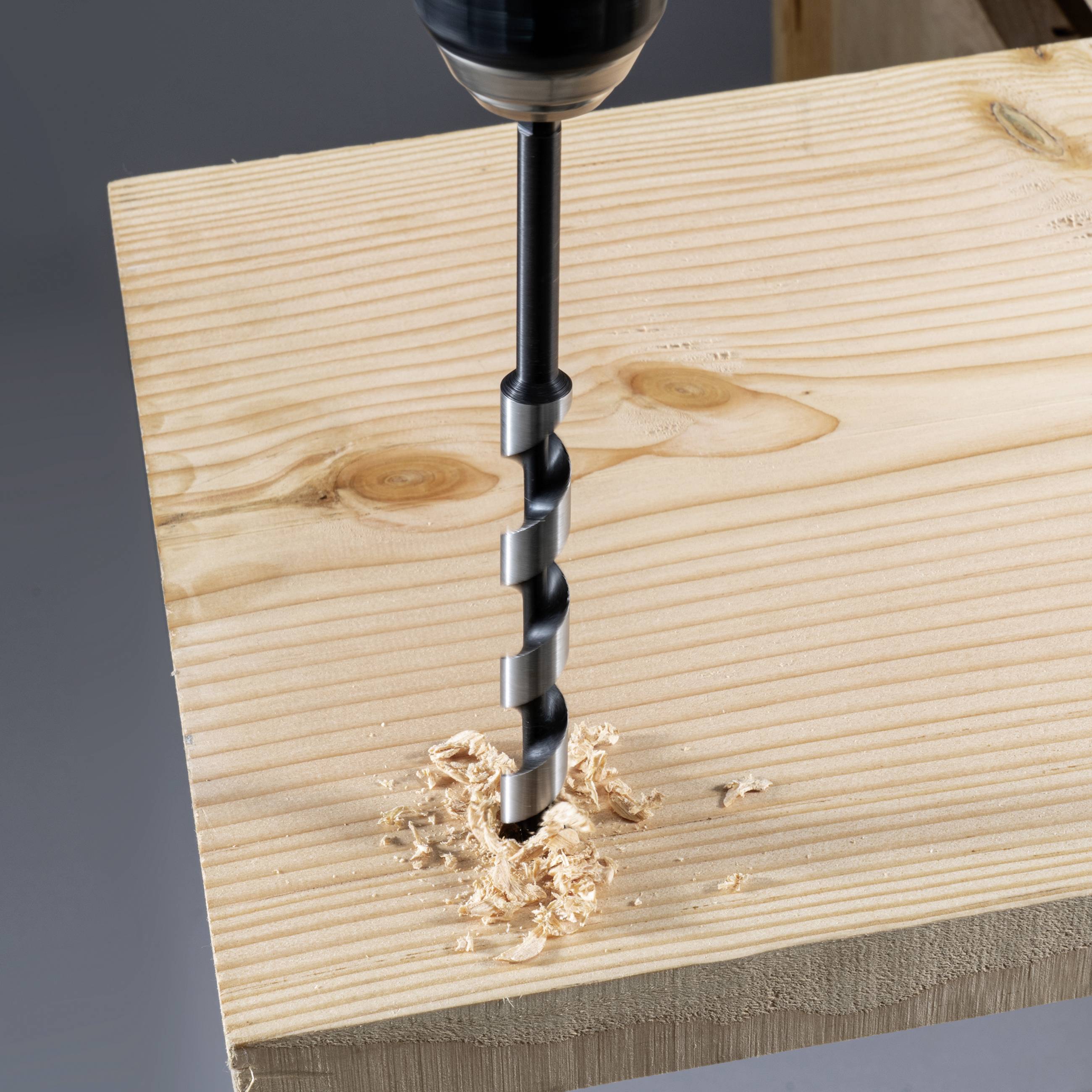 Ein Bohrer durchbohrt ein Holzbrett, Späne sammeln sich um das Loch. Der Fokus liegt auf der Bohrspitze und dem durchtrennten Holz.