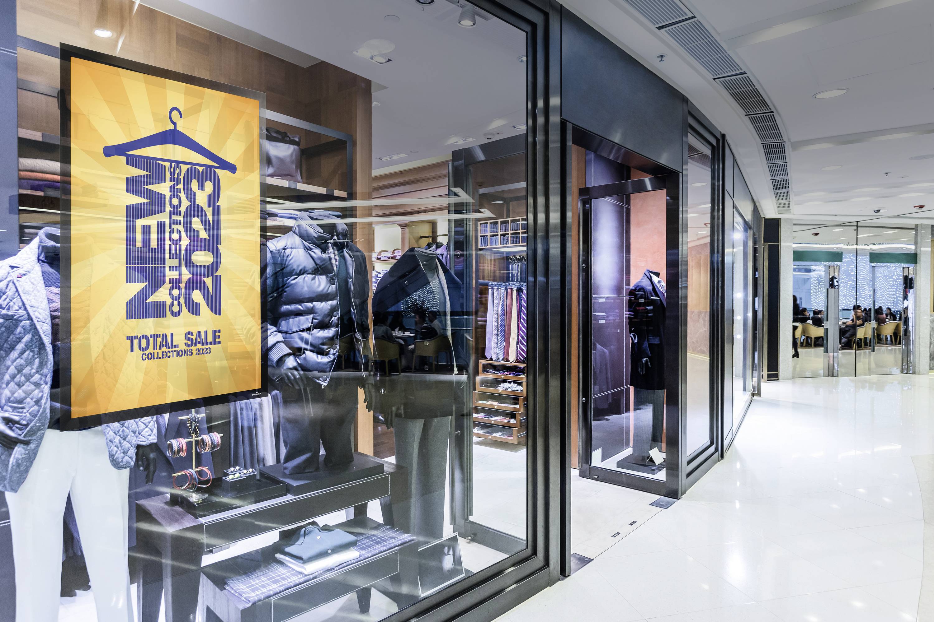 Geschäftsfront mit Schaufenster, das Bekleidung zeigt. Ein Plakat bewirbt 'New Collection 2023 Total Sale'. Lang Flur im Einkaufszentrum.