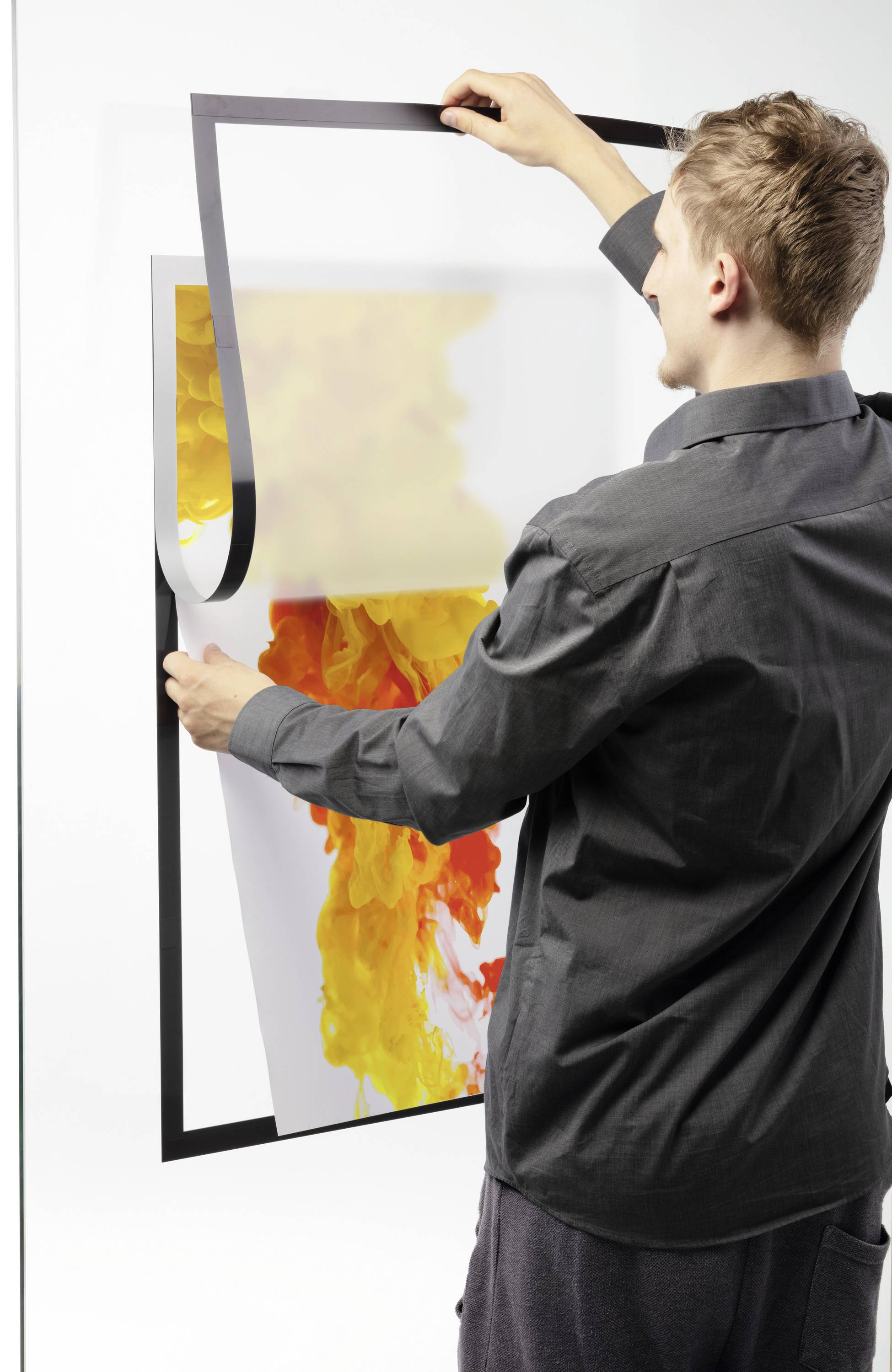 Mann hängt ein buntes Bild mit gelb-orangefarbenem Motiv in einen schwarzen Rahmen an einer weißen Wand auf.