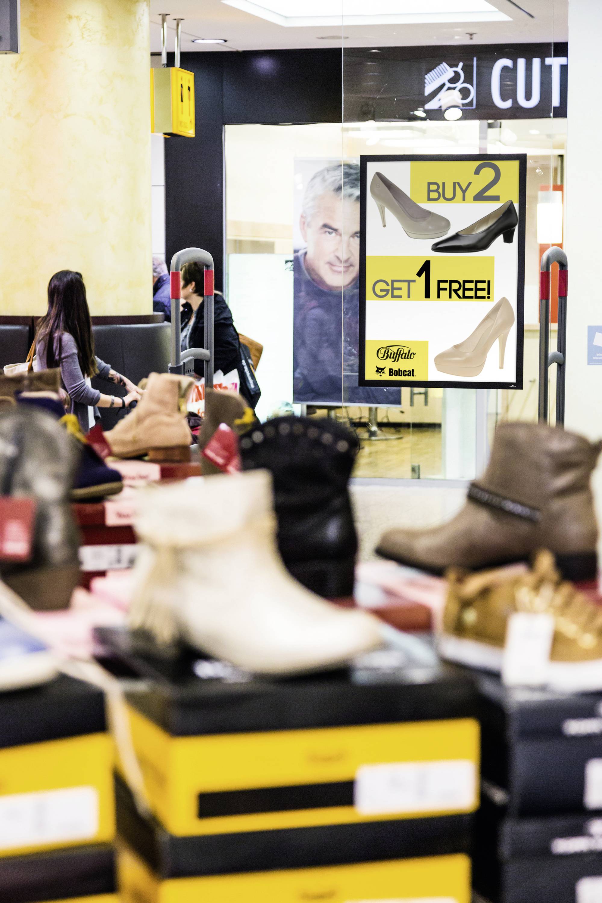 Ein Schuhgeschäft mit einem Werbeplakat im Hintergrund: 'Buy 2, Get 1 Free'. Mehrere Schuhe auf Tischen im Vordergrund.