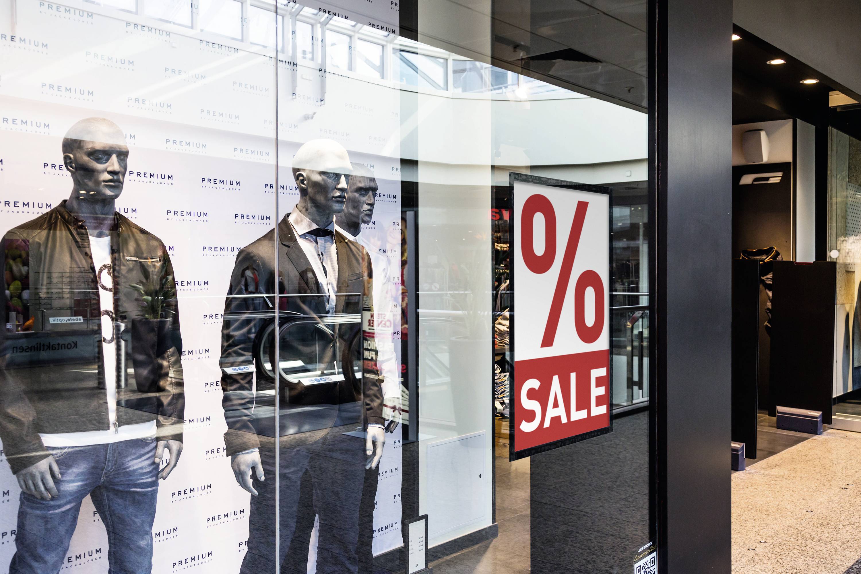 Schaufenster eines Bekleidungsgeschäfts mit zwei Schaufensterpuppen in schwarzen Jacken. Ein großes Schild zeigt '% SALE'.