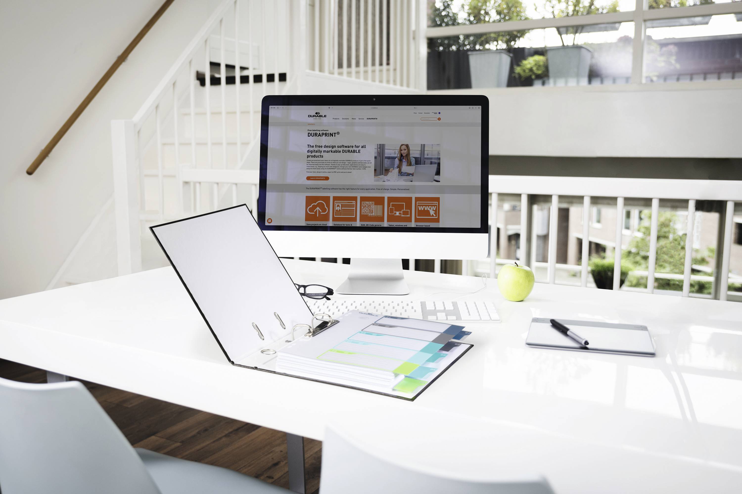 Ein aufgeräumter Schreibtisch mit Computer, geöffnetem Ordner mit Reitern, Notizblock, Stift und Apfel. Große Fenster im Hintergrund.