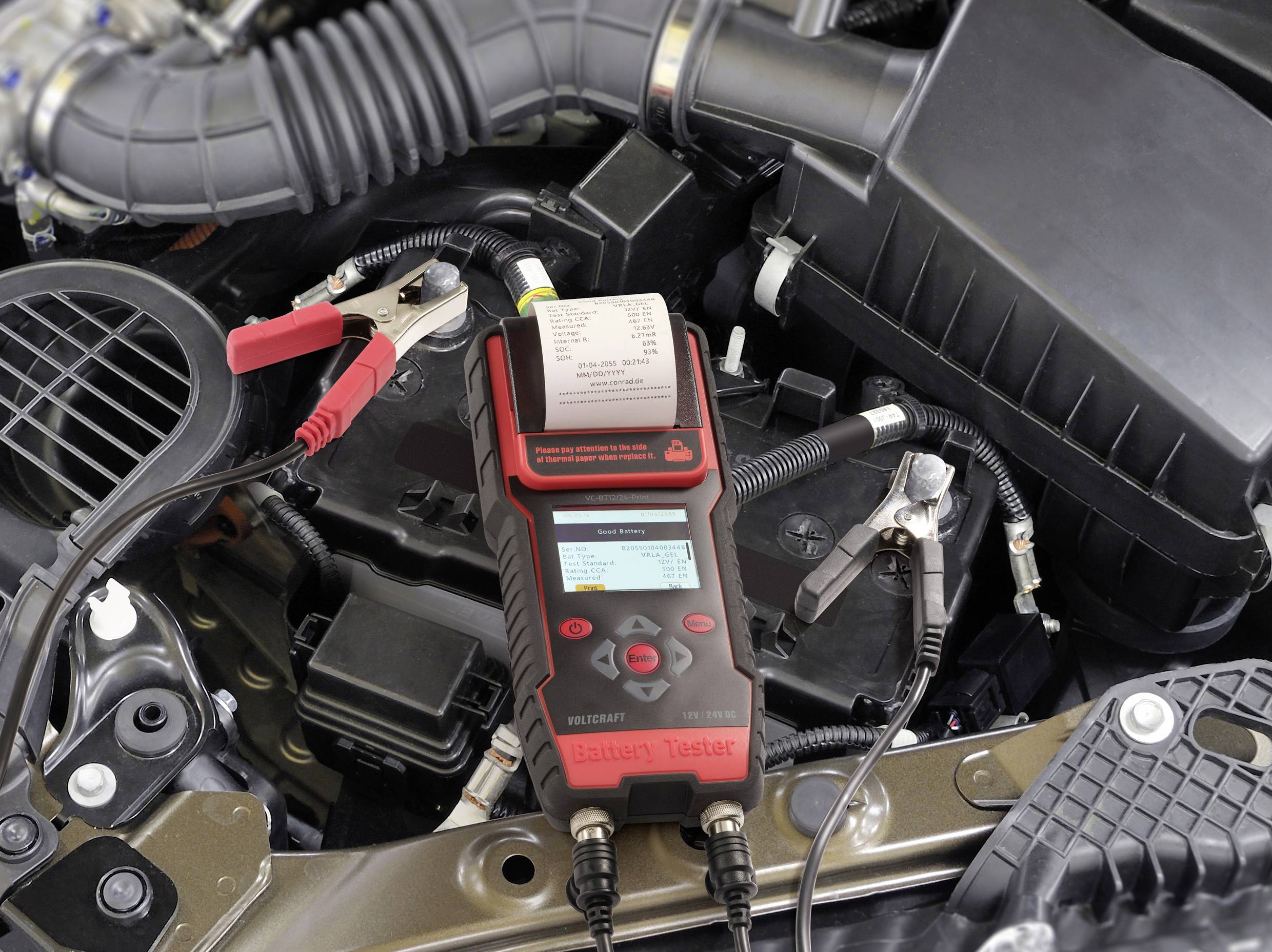 Ein Batterietester ist an eine Autobatterie im Motorraum angeschlossen und zeigt Messdaten auf dem Display an.
