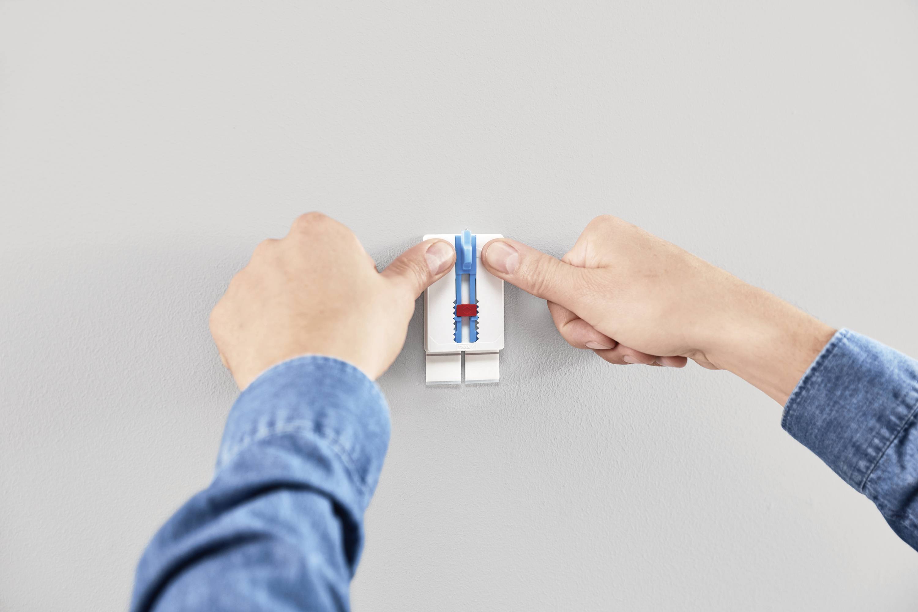 Zwei Hände drücken einen Klebehaken an eine Wand. Der Haken hat einen blauen Klebestreifen.