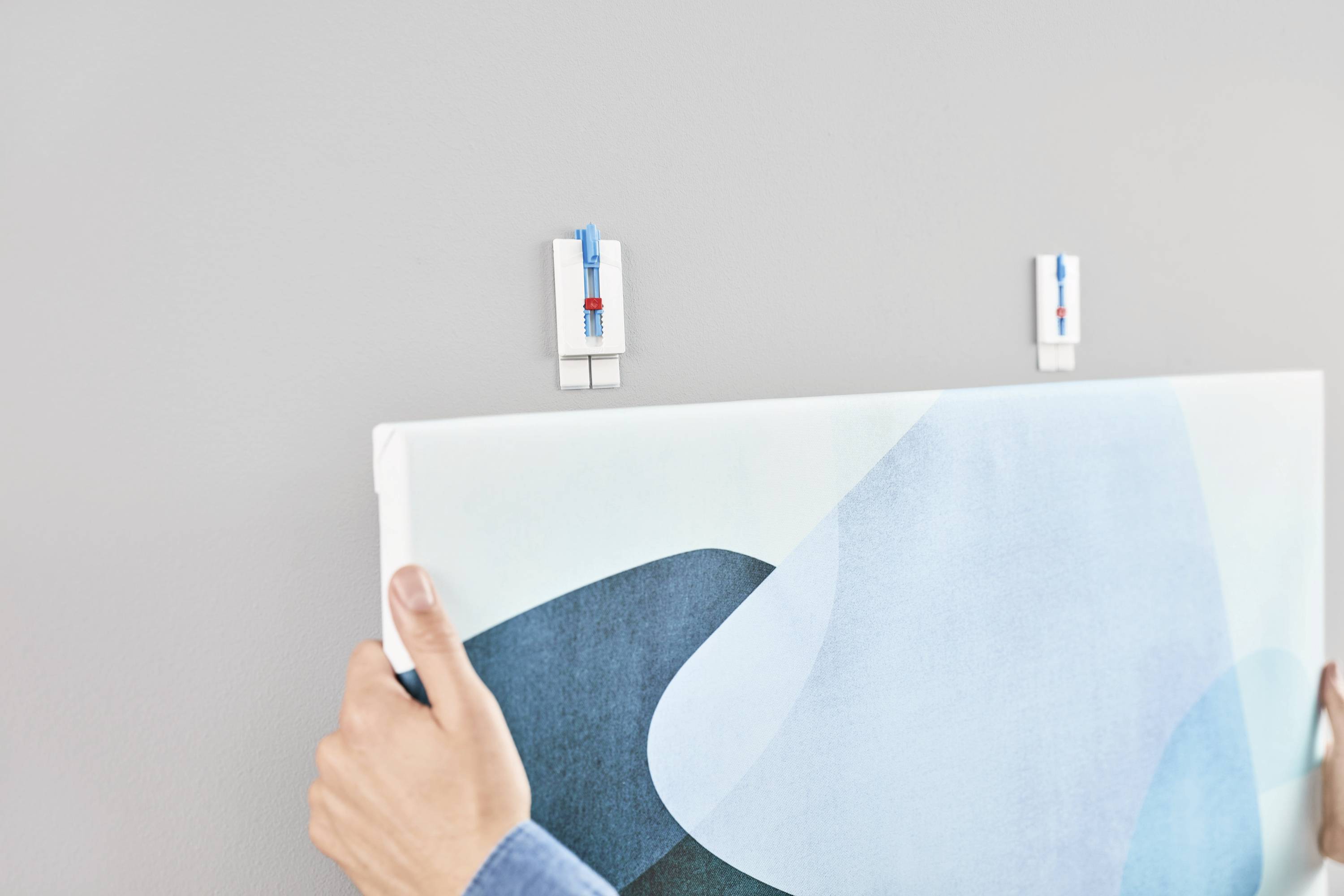 Person hängt ein Bild an die Wand. Bild wird mit zwei weißen Befestigungsklammern fixiert. Hände einer Person sind sichtbar.