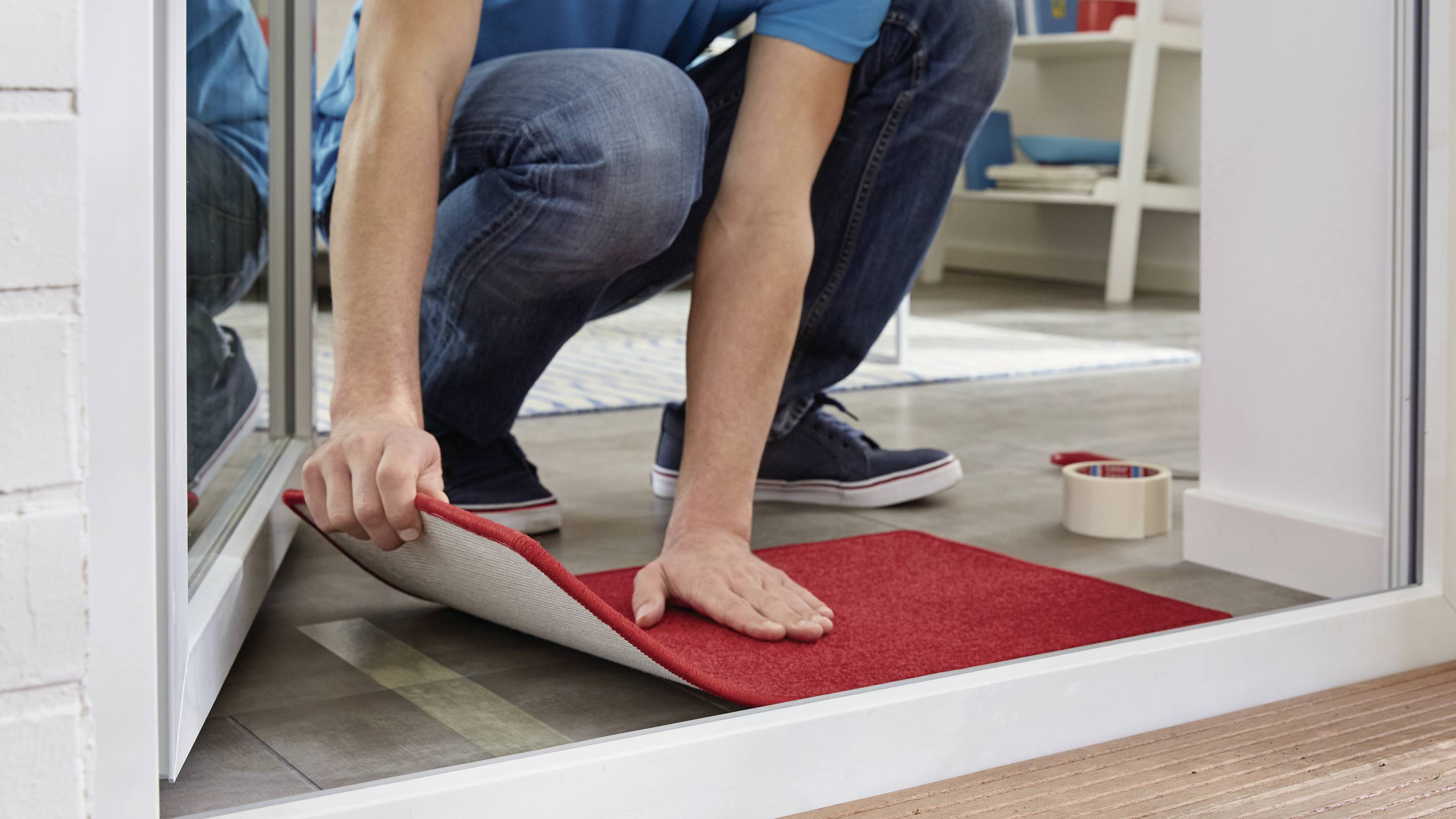 Ein Mann legt einen roten Teppichstreifen auf einen Boden in der Nähe einer Glastür. Eine Rolle Klebeband liegt daneben.
