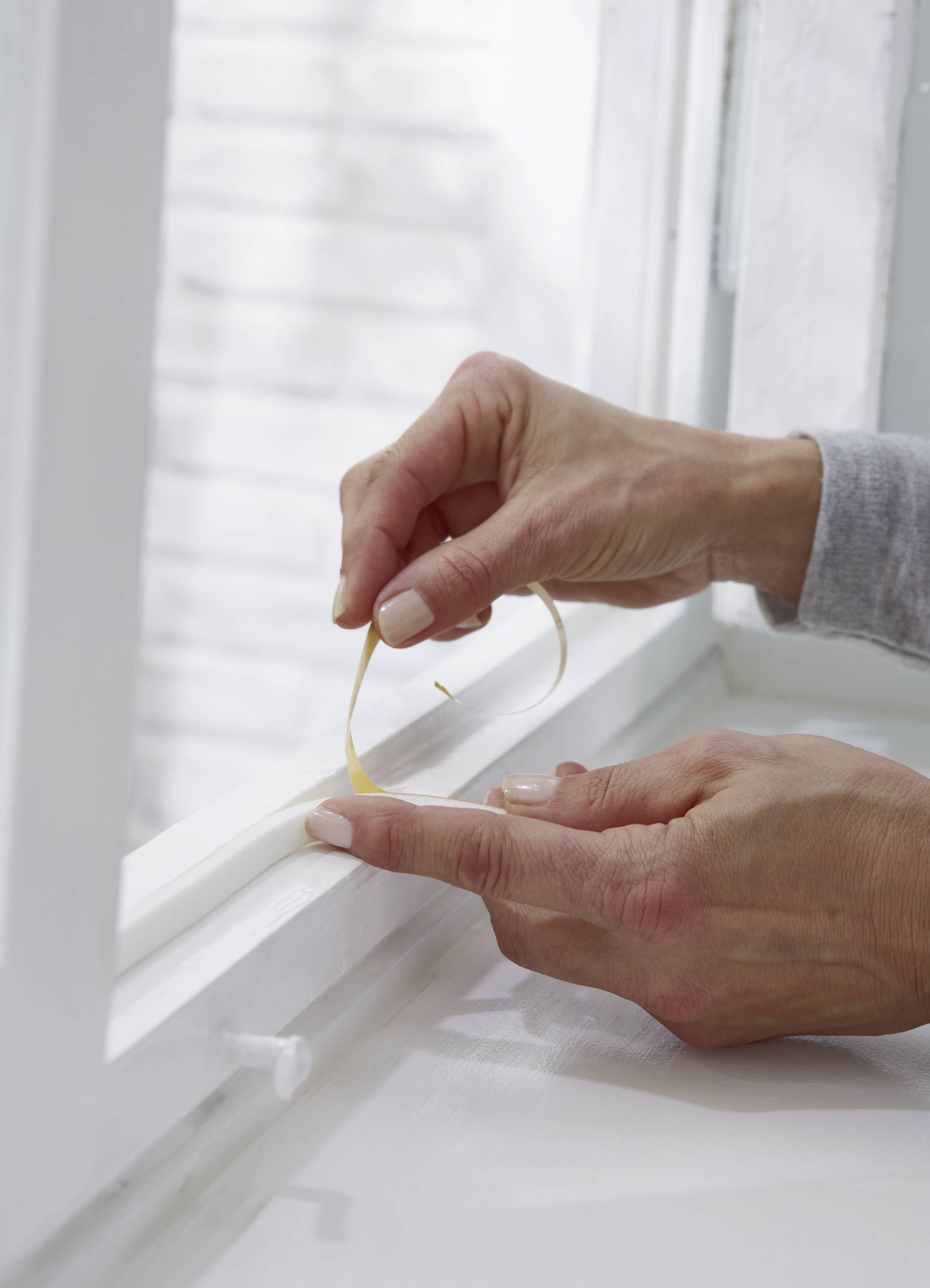 Zwei Hände befestigen ein dichtendes Klebeband an einem Fensterrahmen, um Zugluft zu verhindern.