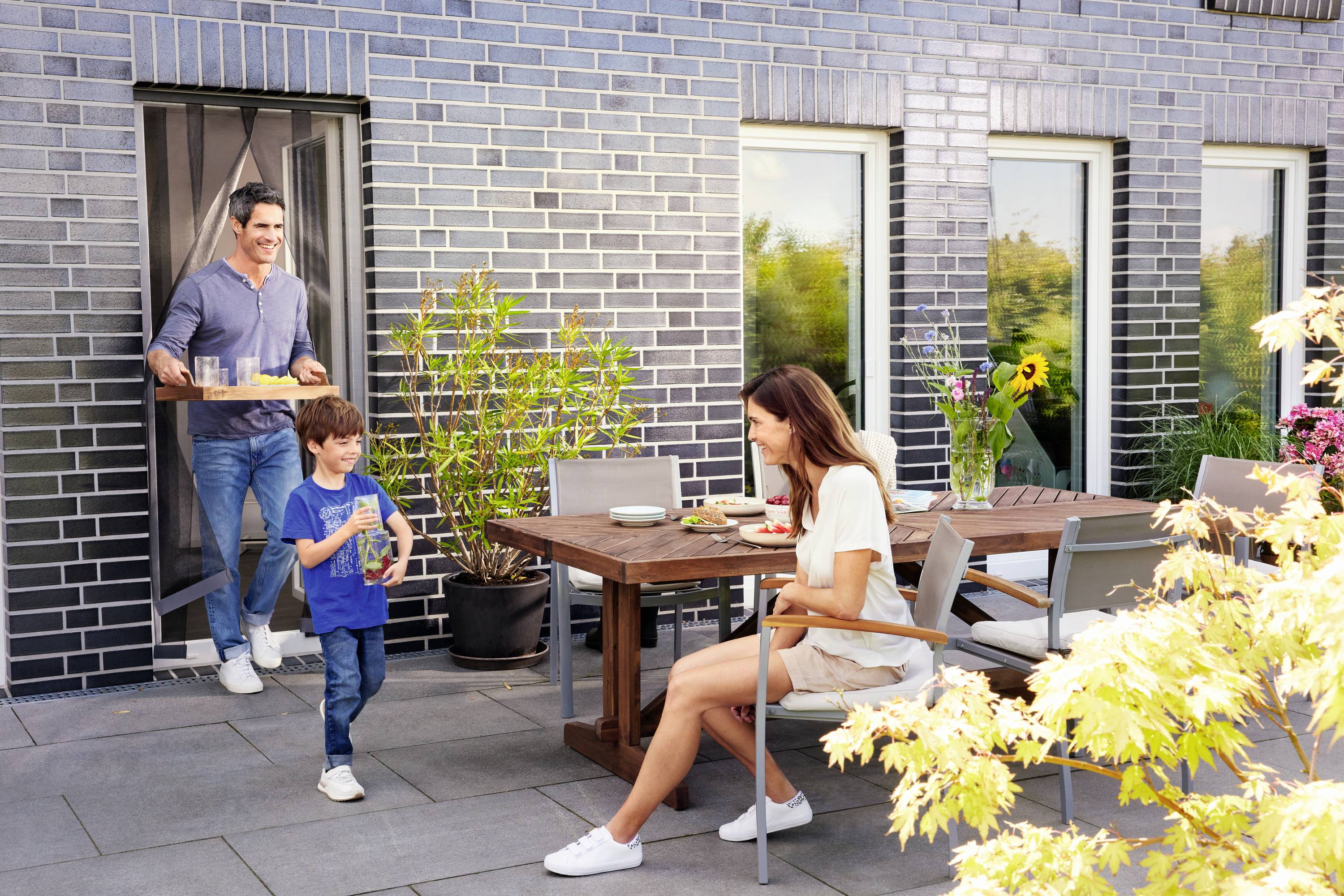 Eine Familie isst auf einer Terrasse. Der Vater bringt ein Tablett, der Sohn hält ein Glas, und die Mutter sitzt am Tisch.