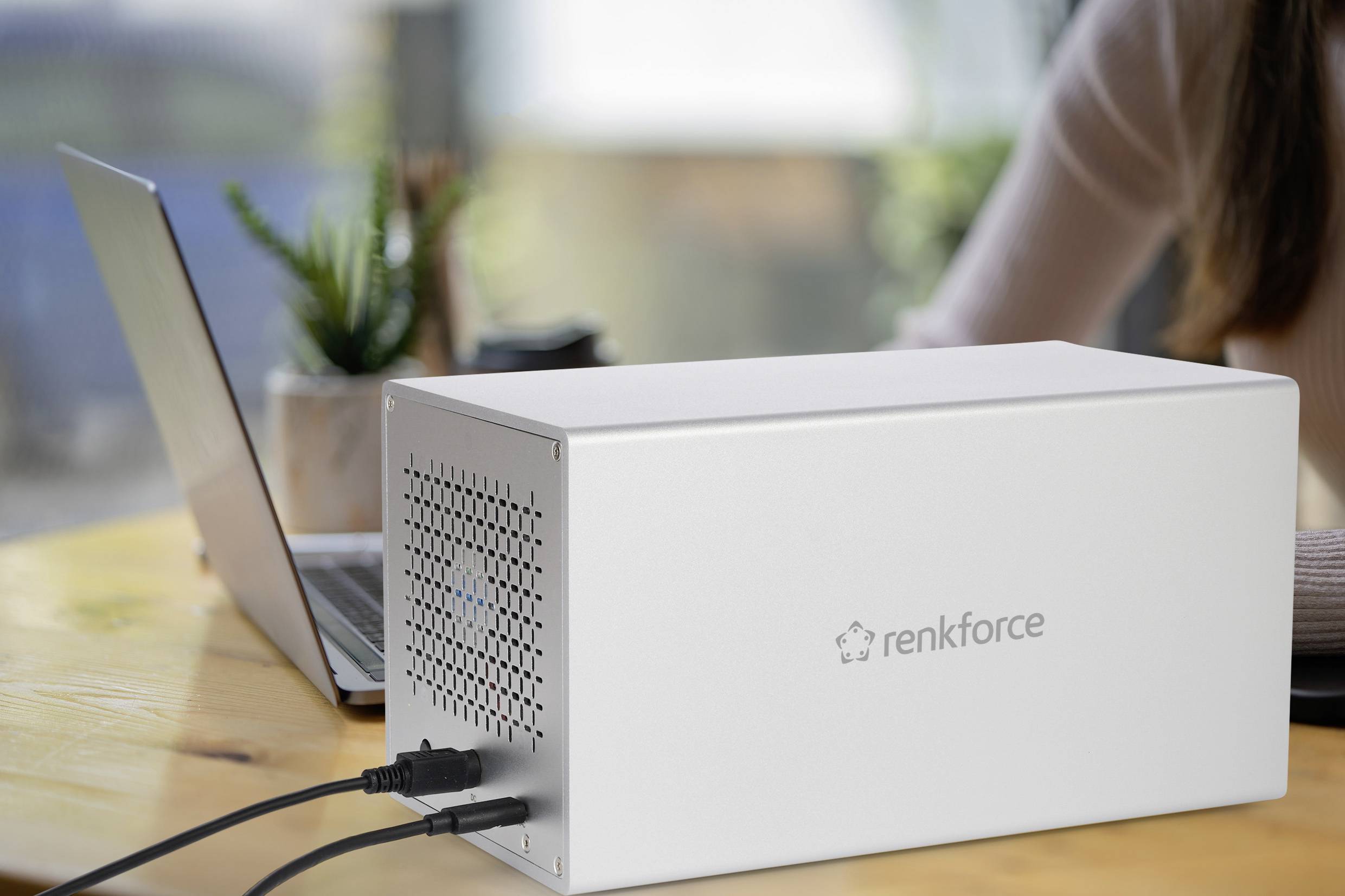 Silbernes 'renkforce'-Gehäuse auf einem Tisch neben einem geöffneten Laptop. Mehrere Kabel sind an das Gehäuse angeschlossen.