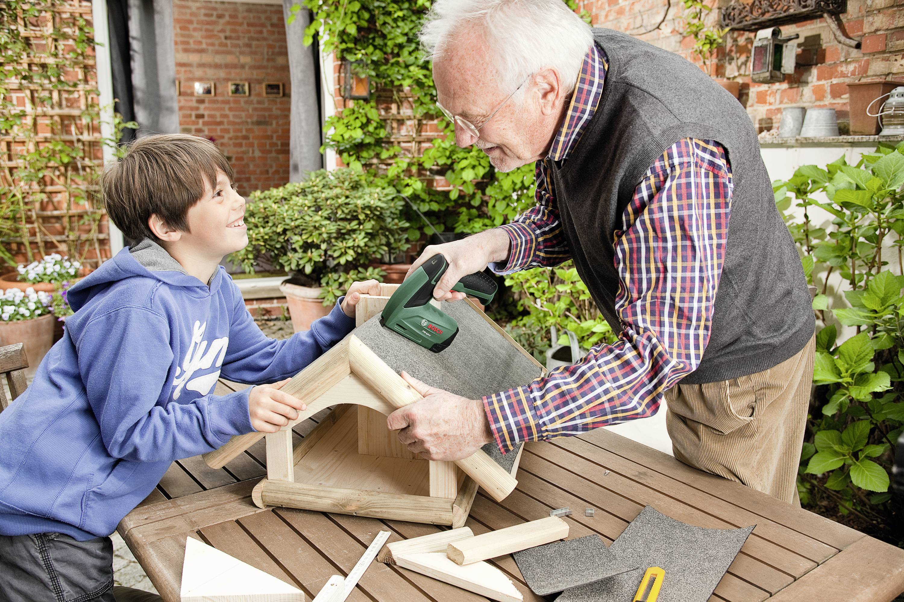 Ein älterer Mann und ein Junge bauen gemeinsam ein Vogelhaus auf einem Holztisch im Garten. Der Mann benutzt ein Werkzeug, der Junge hält Holzteile.
