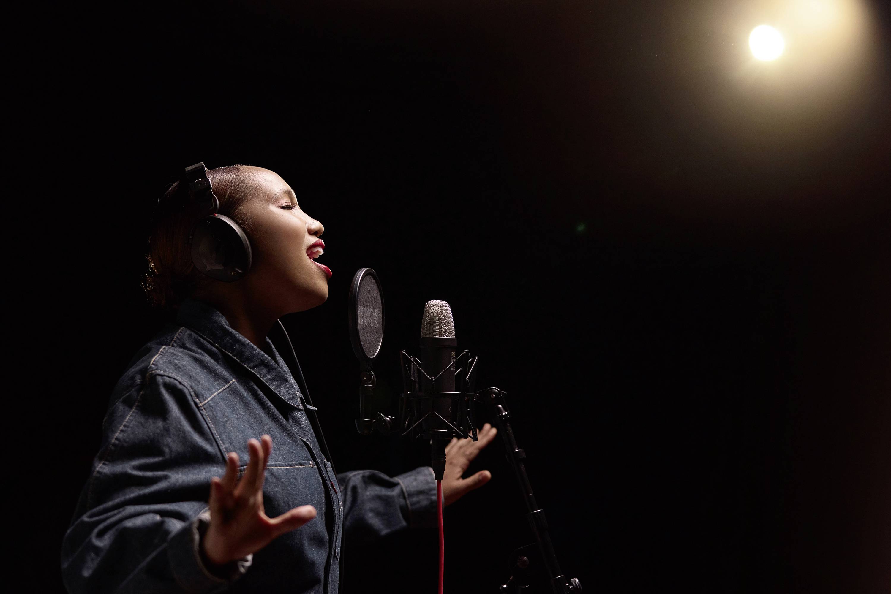 Eine Person mit Kopfhörern singt leidenschaftlich in ein Mikrofon unter einem hellen Scheinwerferlicht in einem Aufnahmestudio.