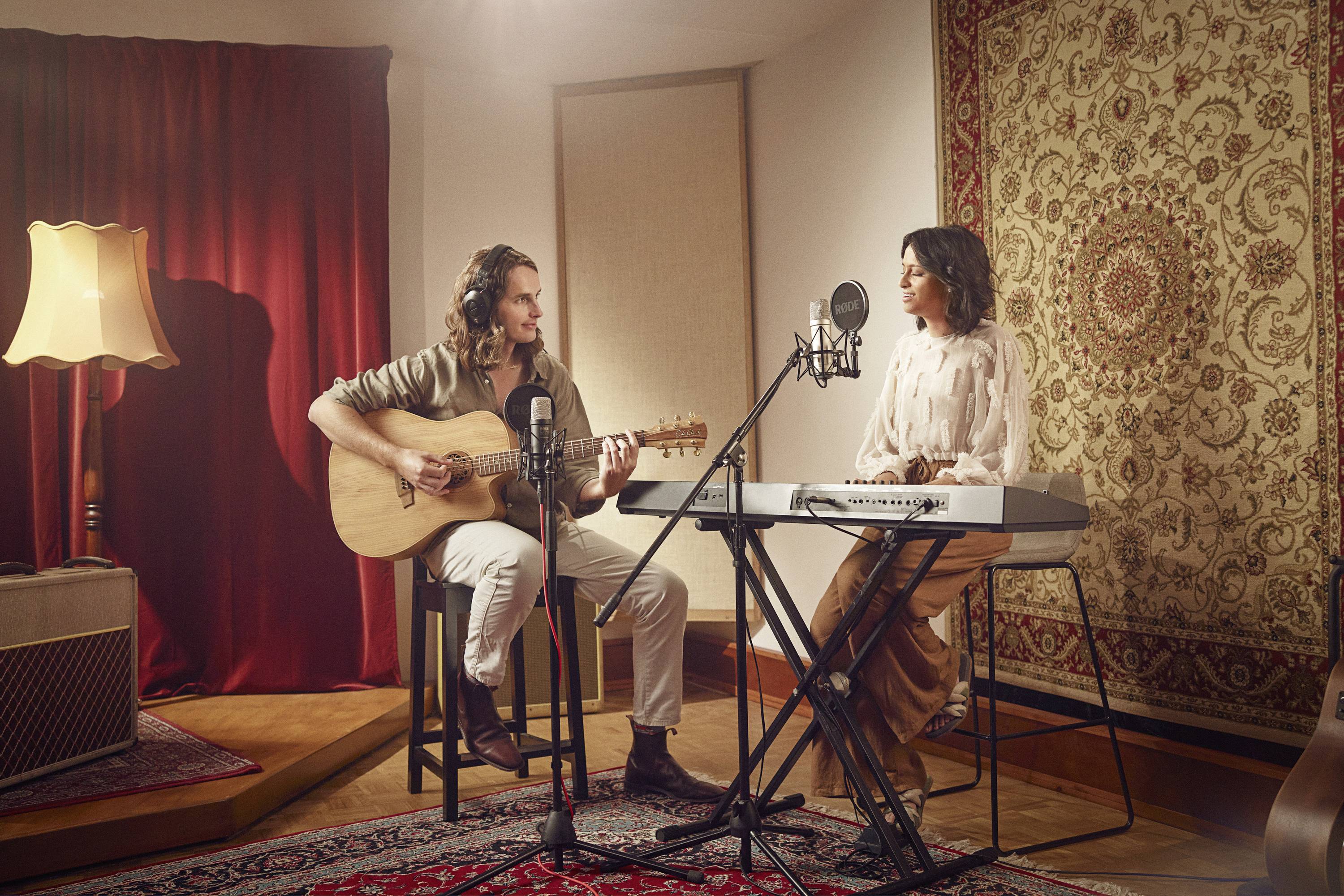 Zwei Personen machen Musik in einem Studio. Links spielt jemand Gitarre, rechts spielt jemand ein Keyboard. Mikrofone sind aufgestellt.