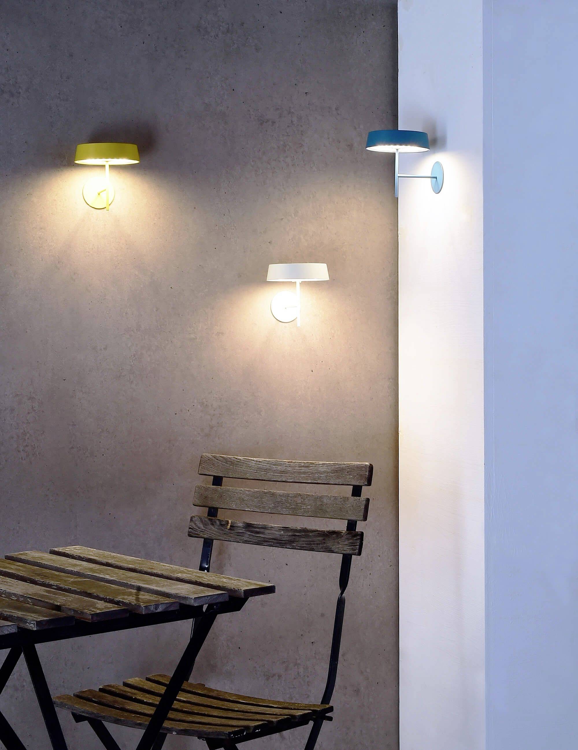 Drei moderne Wandleuchten in Grün, Weiß und Blau erhellen eine graue Wand über einem klappbaren Holztisch und Stuhl.