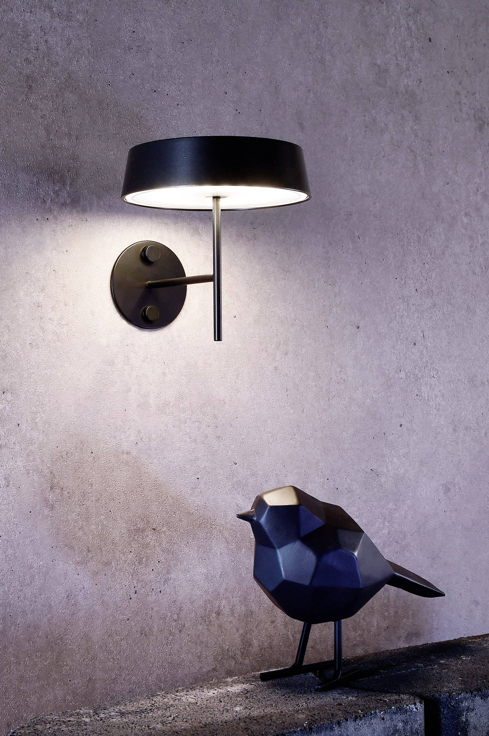 Eine minimalistische schwarze Wandleuchte leuchtet eine geometrische Vogelfigur aus Metall auf einem Betonregal an einer grauen Wand aus.
