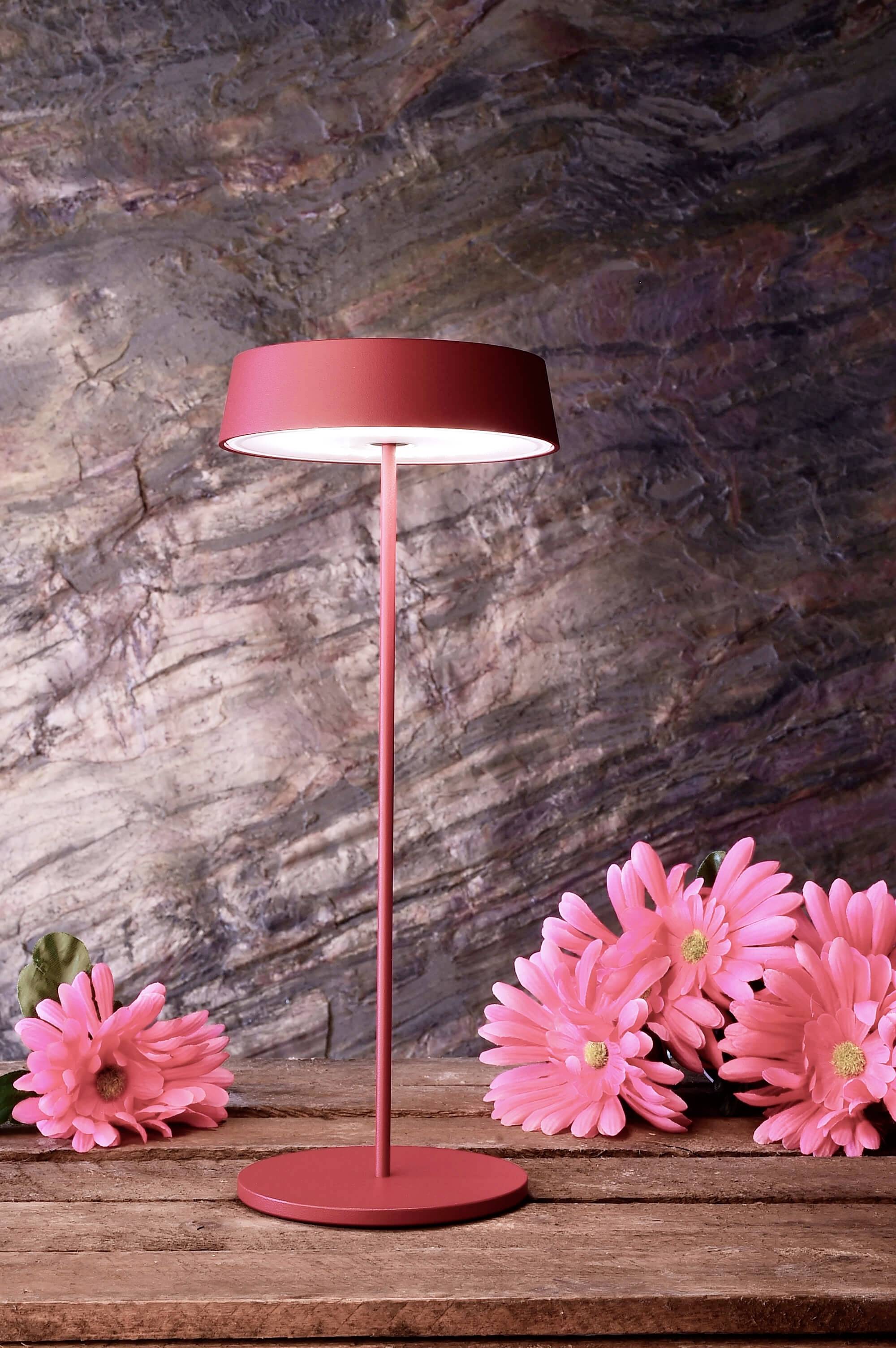 Eine rote Tischlampe leuchtet auf einem Holztisch. Daneben liegen mehrere rosa Gerbera-Blumen. Der Hintergrund ist steinartig.