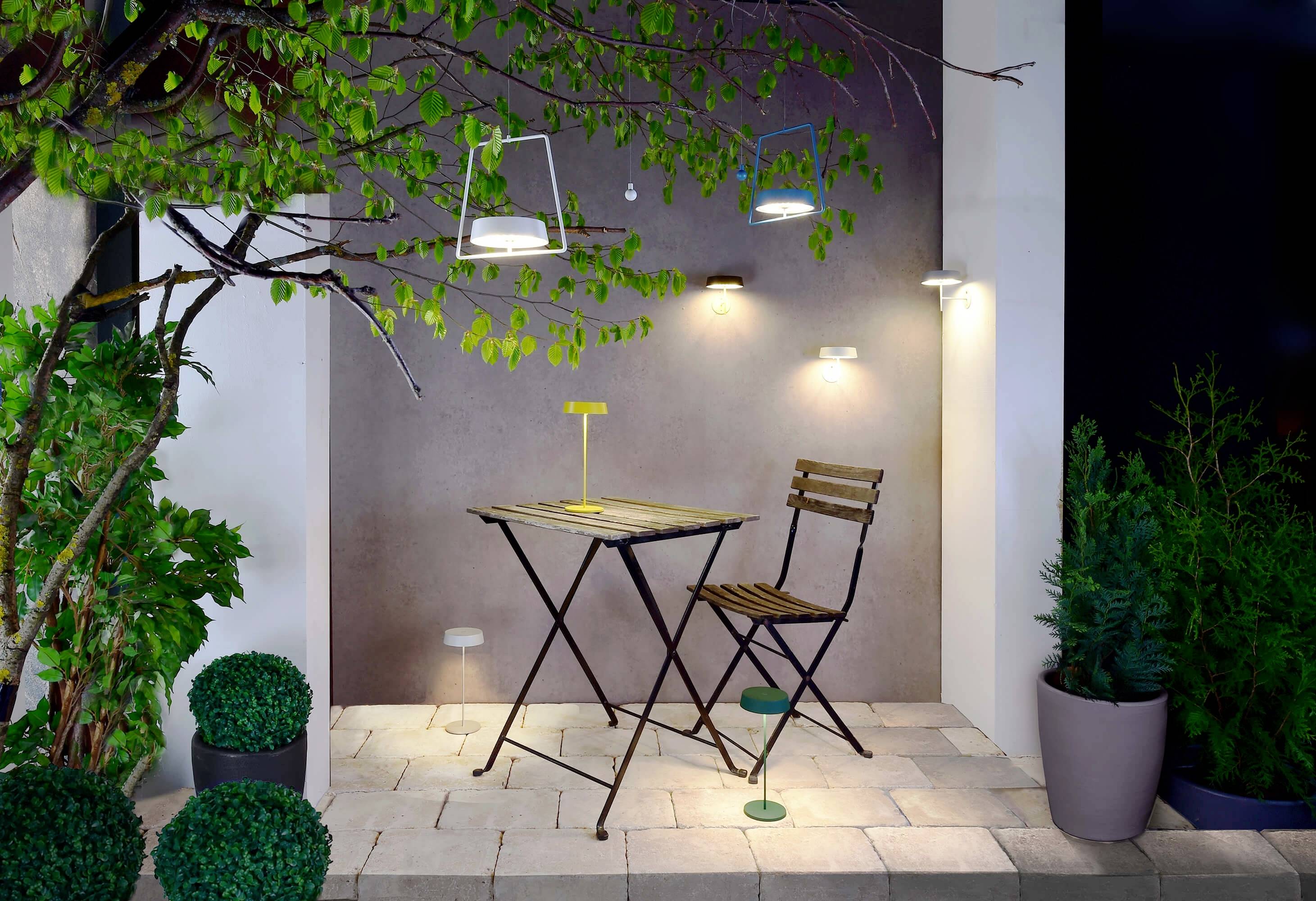 Ein kleiner Balkon mit einem Tisch und einem Stuhl. Umgeben von Pflanzen, darunter ein Baum rechts und Kübelpflanzen links. Beleuchtung an der Wand.