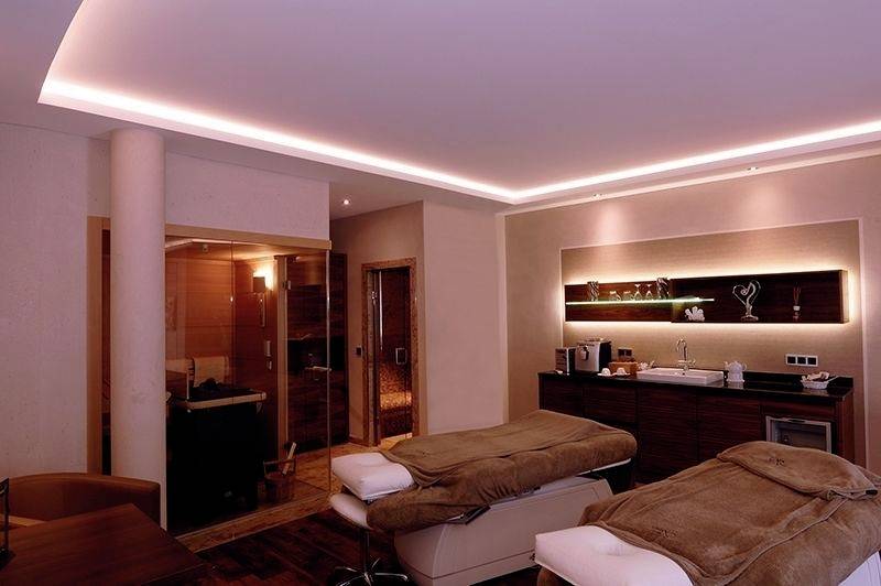 Zwei Massageliegen in einem Spa-Raum mit gedimmter Beleuchtung, hellen Wänden und Holzdekor. Im Hintergrund ist eine Sauna zu sehen.