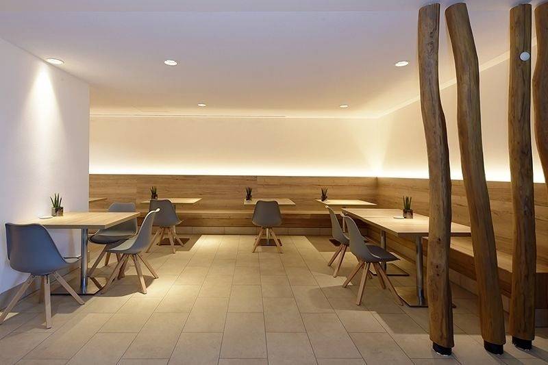 Ein moderner, minimalistischer Speiseraum mit Holztischen und -bänken, vereinzelten Pflanzen als Dekoration, beleuchtet durch dezentes, warmes Licht.
