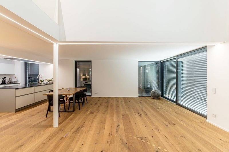 Großer, moderner Wohnraum mit Holzboden, Esstisch mit Stühlen rechts und Küche links. Große Fensterfronten mit Jalousien.
