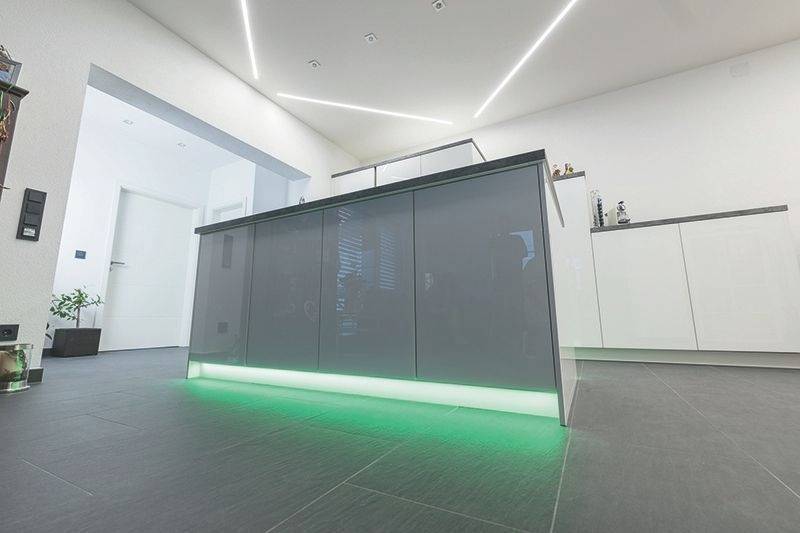 Küche mit moderner grauer Insel, darunter grüne Beleuchtung. Weiße Wände, minimalistisches Design, hell erleuchtet.