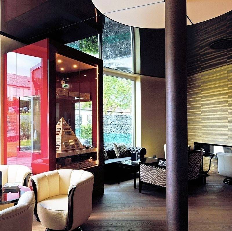 Ein modernes Café mit stilvollen Sesseln, großen Fenstern und einer Ausstellungsvitrine. Gemütliche Atmosphäre und natürliche Beleuchtung.