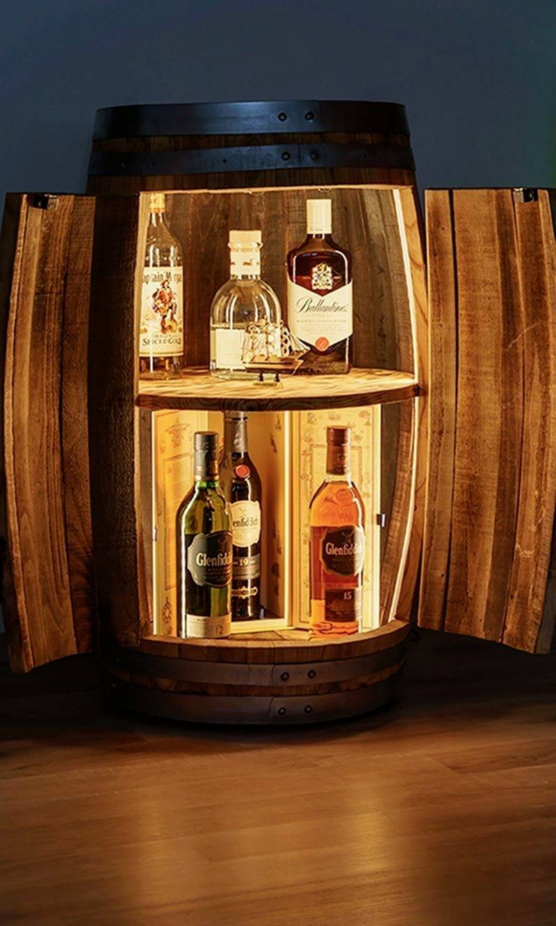 Holzfass mit offener Tür zeigt verschiedene Flaschen Alkohol, darunter Whisky und Gin, auf einem Regal im Inneren, beleuchtet von warmem Licht.