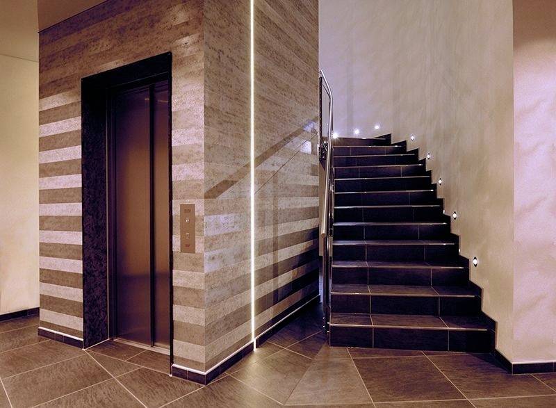 Modernes Treppenhaus mit Lift; rechte Treppe aus dunklem Stein mit eingelassenen Lichtern, links metallischer Aufzug mit Holzverkleidung.