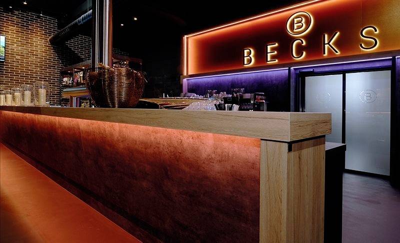 Barbereich mit einem Holz- und Metalltresen, im Hintergrund ein Leuchtschild mit der Aufschrift 'BECKS'.