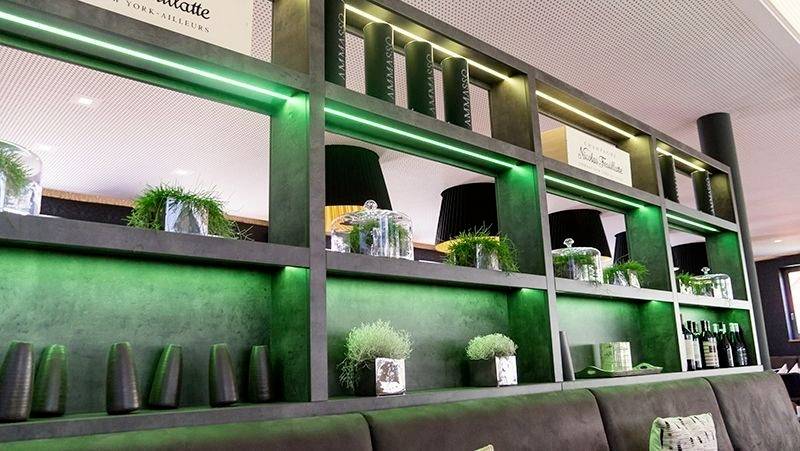 Eine moderne Bar mit grüner LED-Beleuchtung, dekoriert mit Glasgefäßen und Pflanzen. Schwarze Regale und Sitzgelegenheiten sind sichtbar.