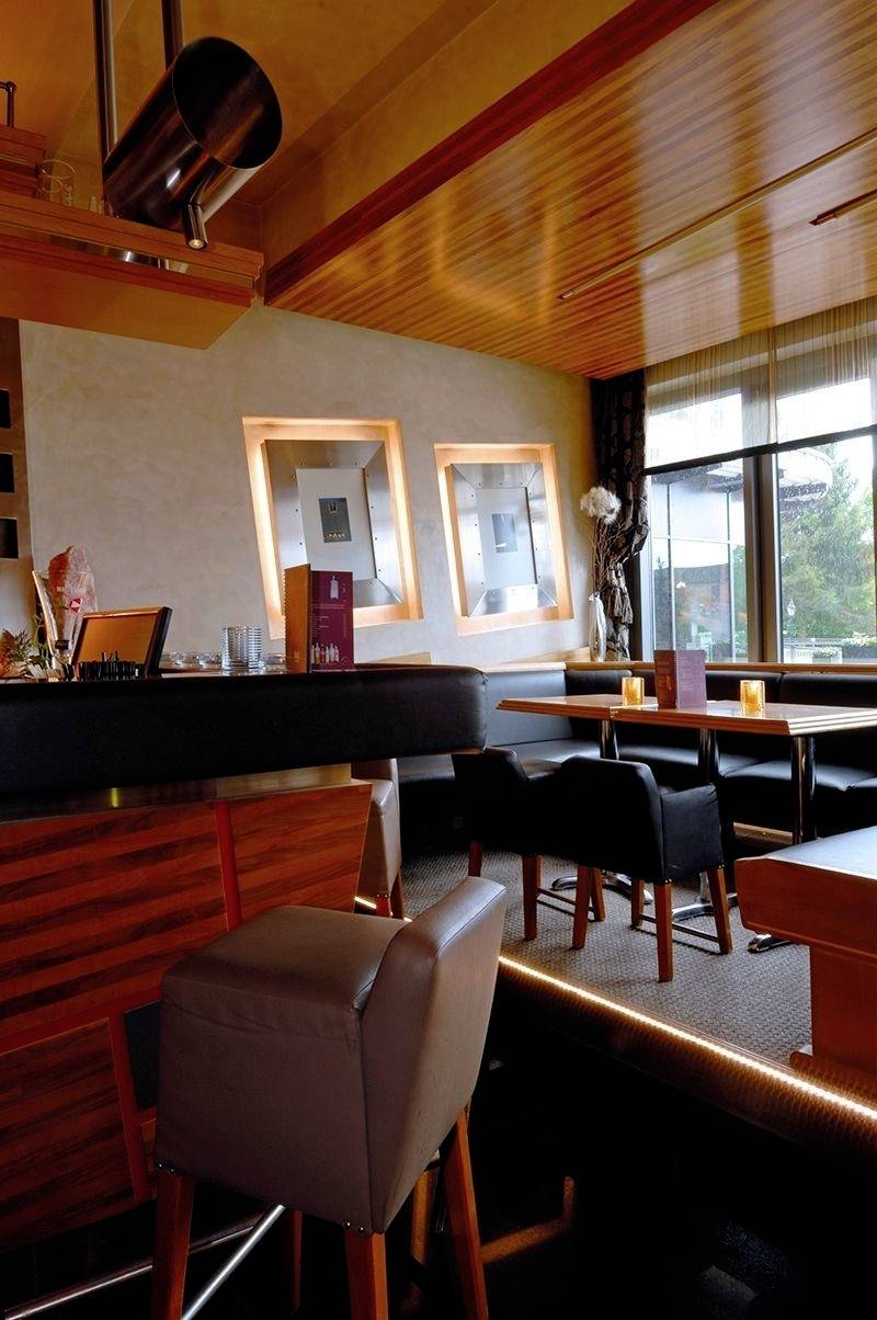 Gemütliches Café mit Holzmöbeln, Barhockern und Tischen, gedämpftem Licht und großen Fenstern, die eine Aussicht auf Bäume bieten.