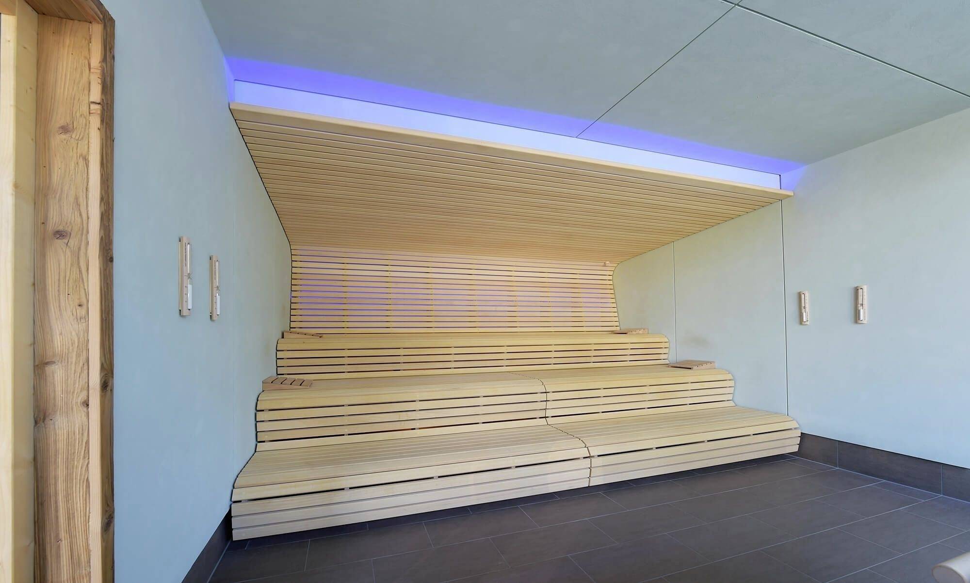 Innenansicht einer modernen Sauna mit hölzernen Sitzbänken, blauer Beleuchtung und hellem Holzwanddesign.