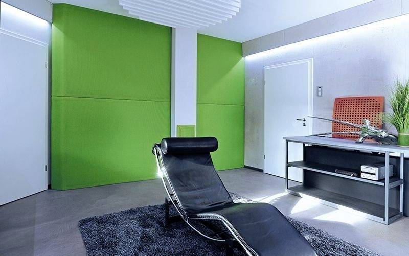 Ein modernes Zimmer mit einem schwarzen Liegesessel auf einem dunklen Teppich, grüne Wandverkleidung, Tisch rechts, Wandregal.