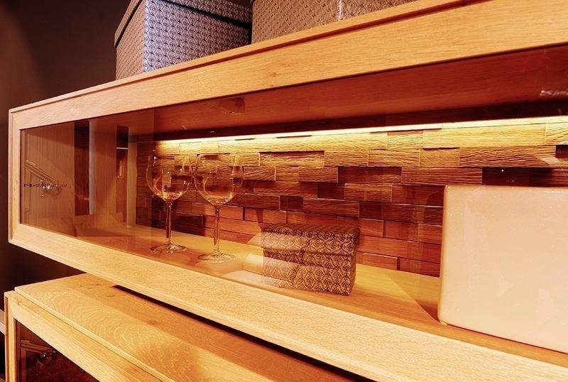 Ein Holzregal mit beleuchtetem Fach zeigt zwei Weingläser und dekorative Boxen. Warmes Licht betont die hölzerne Rückwand.