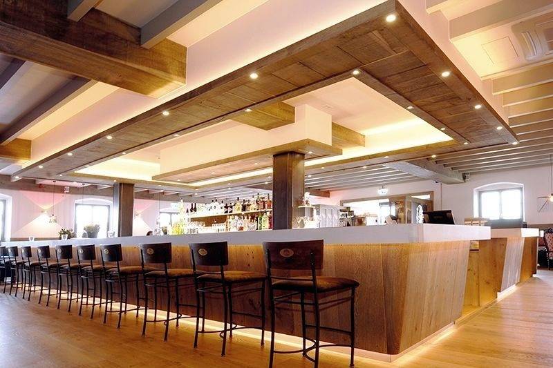 Moderne Bar mit Holzdekor und beleuchteter Theke. Mehrere Barhocker stehen in einer Reihe. Im Hintergrund sind Flaschenregale sichtbar.
