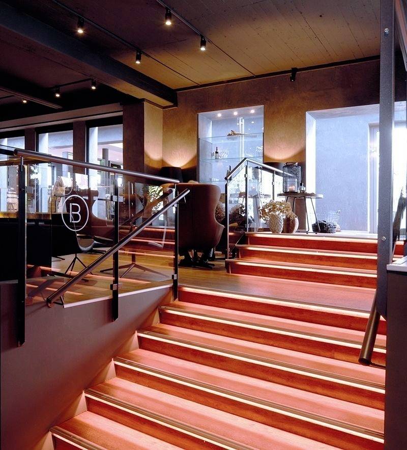 Eine Treppe mit roter Beleuchtung in einem modernen Interieur. Rechts befindet sich ein Tisch mit Dekorationen, links eine Glaswand mit einem Symbol.