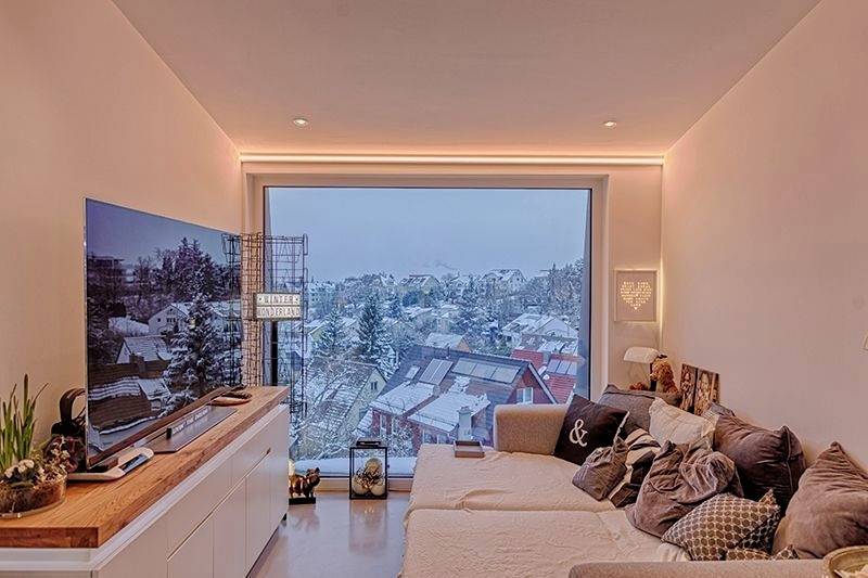 Modernes Wohnzimmer mit beige Sofa, großem Fernseher und Panoramafenster mit Blick auf verschneite Häuser und Bäume.