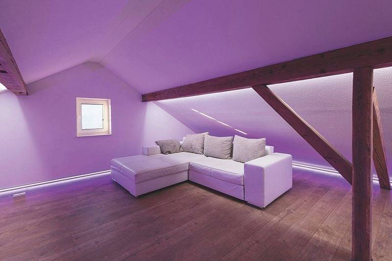 Minimalistisch eingerichtetes Dachgeschosszimmer mit lila Beleuchtung, einem weißen Ecksofa und Holzbalken.