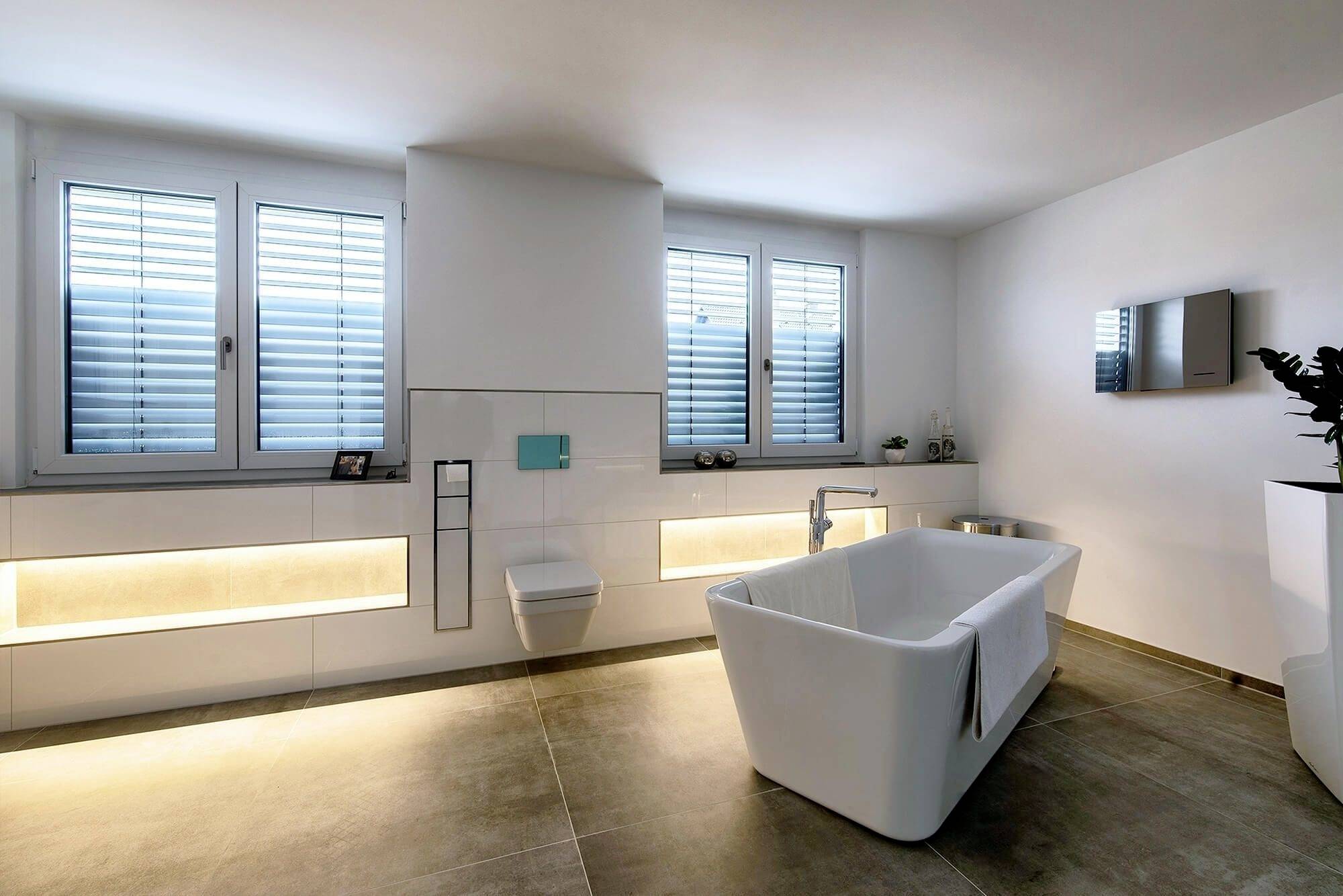 Helles, modernes Badezimmer mit freistehender Badewanne, wandhängendem WC und beleuchteter Ablage unter breiten Fenstern.