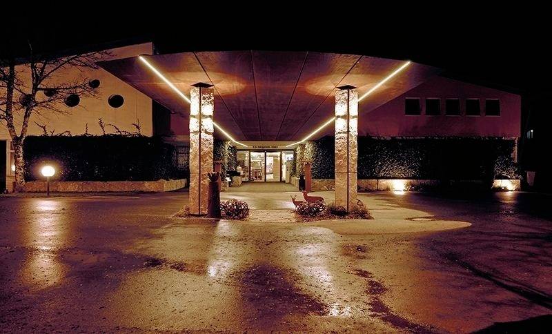 Hotel-Eingang bei Nacht mit beleuchtetem Überdach. Lichtakzente an Säulen führen zum Eingang. Parkplatz im Vordergrund.