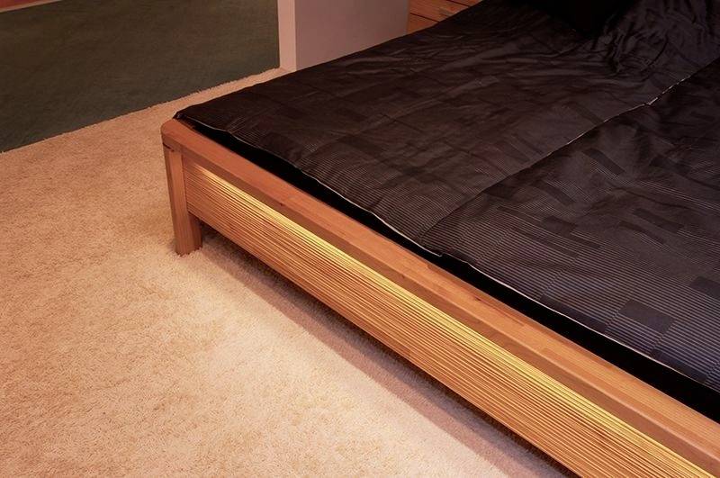 Ein Holzbett mit schwarzer Bettdecke steht auf einem beigen Teppich. Der Boden im Hintergrund ist teils mit dunklem Teppich bedeckt.