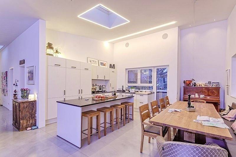 Moderne Küche und Essbereich mit weißem Design, zentralem Inselblock mit Stühlen, großem Esstisch, Klavier und dezentem Deckenspot.