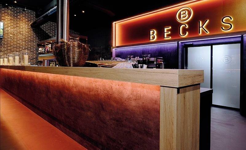 Eine stilvolle Bar mit moderner Beleuchtung. Neonleuchtschild mit der Aufschrift 'BECKS'. Holztheke im Vordergrund; Glasprodukte im Hintergrund.