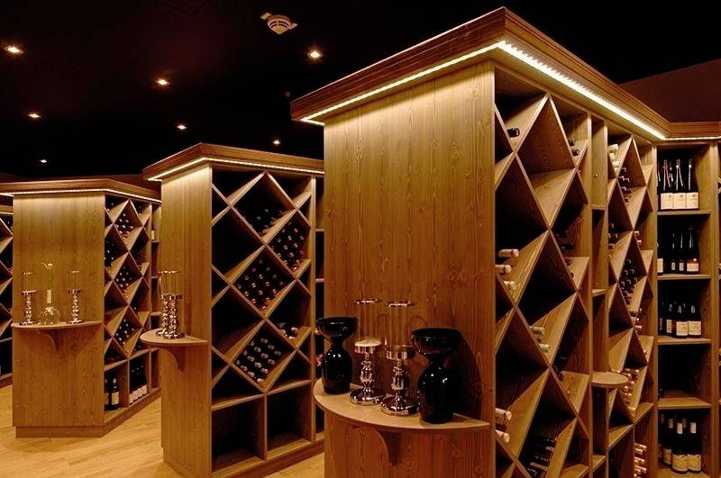 Weinkeller mit Holzeinrichtung, beleuchtet. Regale mit weinbestückten Fächern. Elegante Dekorationen, darunter Kerzenhalter.