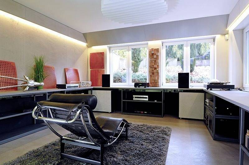 Moderner Raum mit Lederliege, Teppich und Audiogeräten. Große Fenster bieten Aussicht auf Garten. Helmutlich Heimkino- oder Musikzimmer.