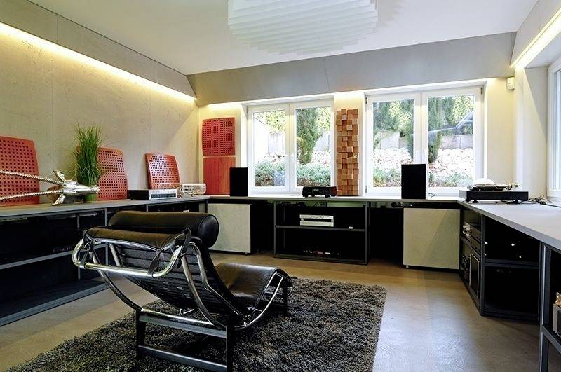 Moderne Lounge mit Liegestuhl, Teppich und Soundsystem. Große Fenster bieten Ausblick auf einen Garten. Elegantes, gemütliches Ambiente.